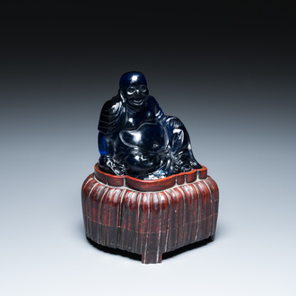 A Chinese blue Beijing glass figure of the laughing Buddha seated on a wooden stand, 19th C.